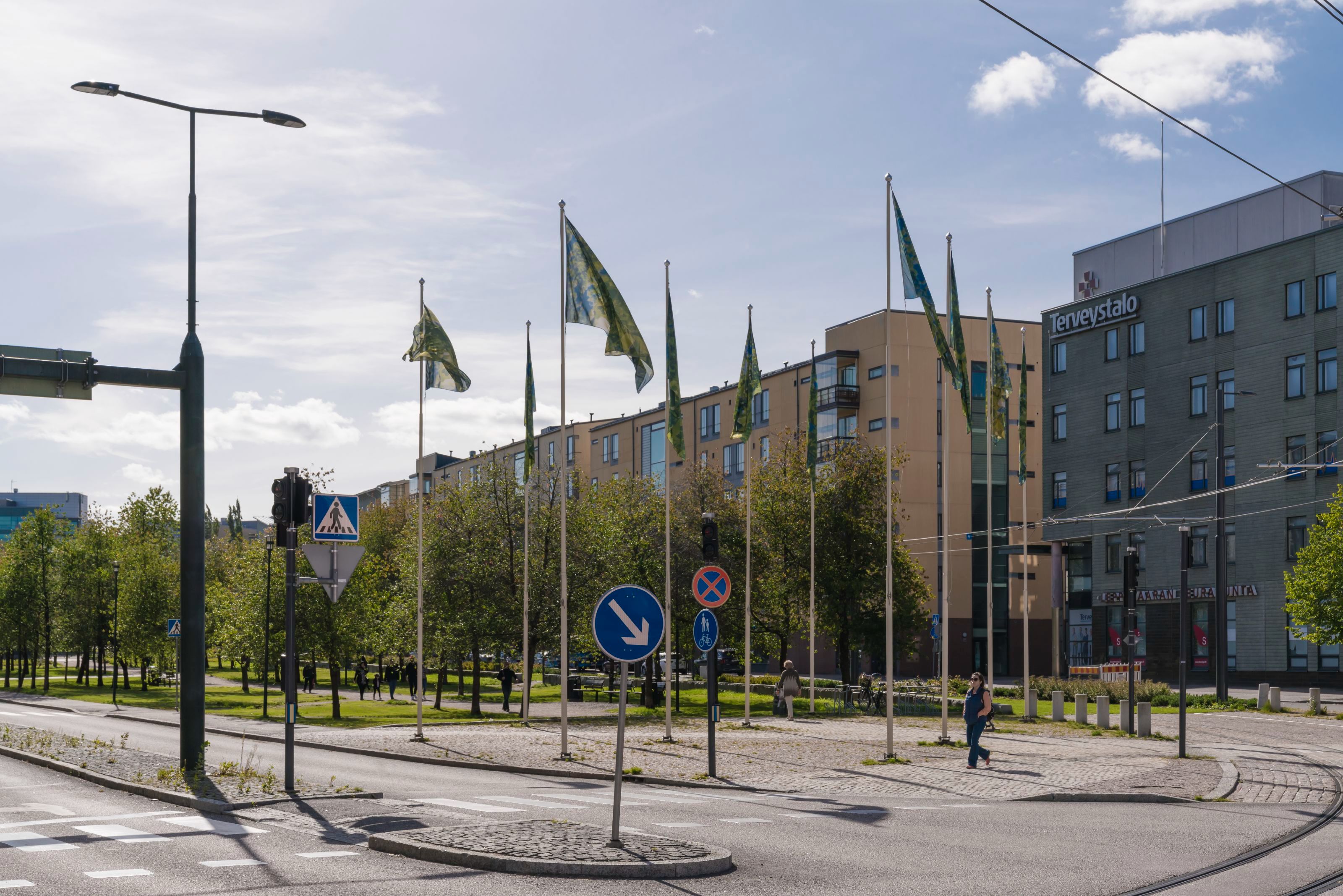 10 lipputankoa, joissa liehuvat liput aurinkoisena päivänä. Taustalla viheralue. Etualalla suojatie ja raitiovaunukiskot.