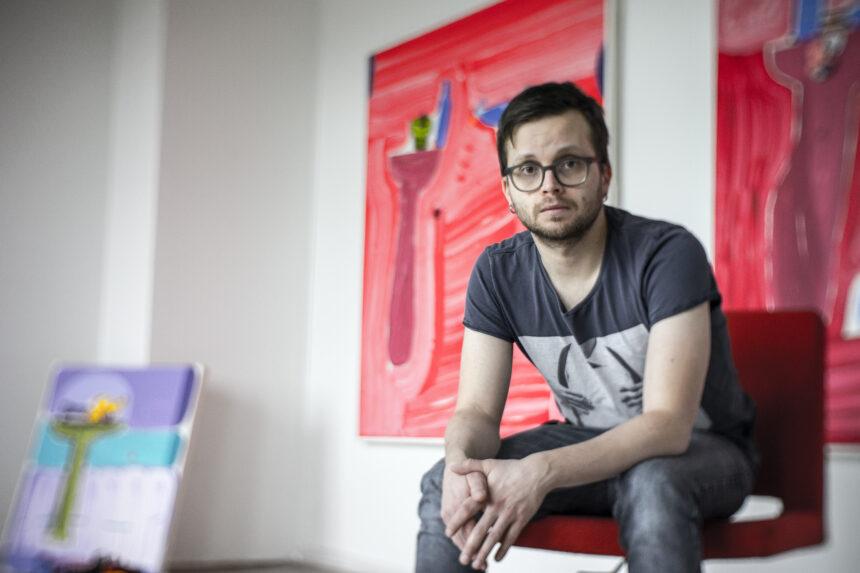 Taiteilija Jussi Goman istuu työhuoneellaan. Taustalla punasävyinen maalaus.