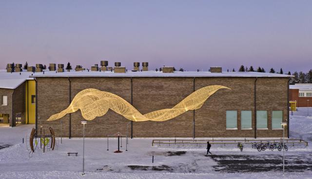 Koulurakennuksen ulkoseinässä suuri kullanvärinen teräksinen taideteos, joka kuvaa veden virtausta.